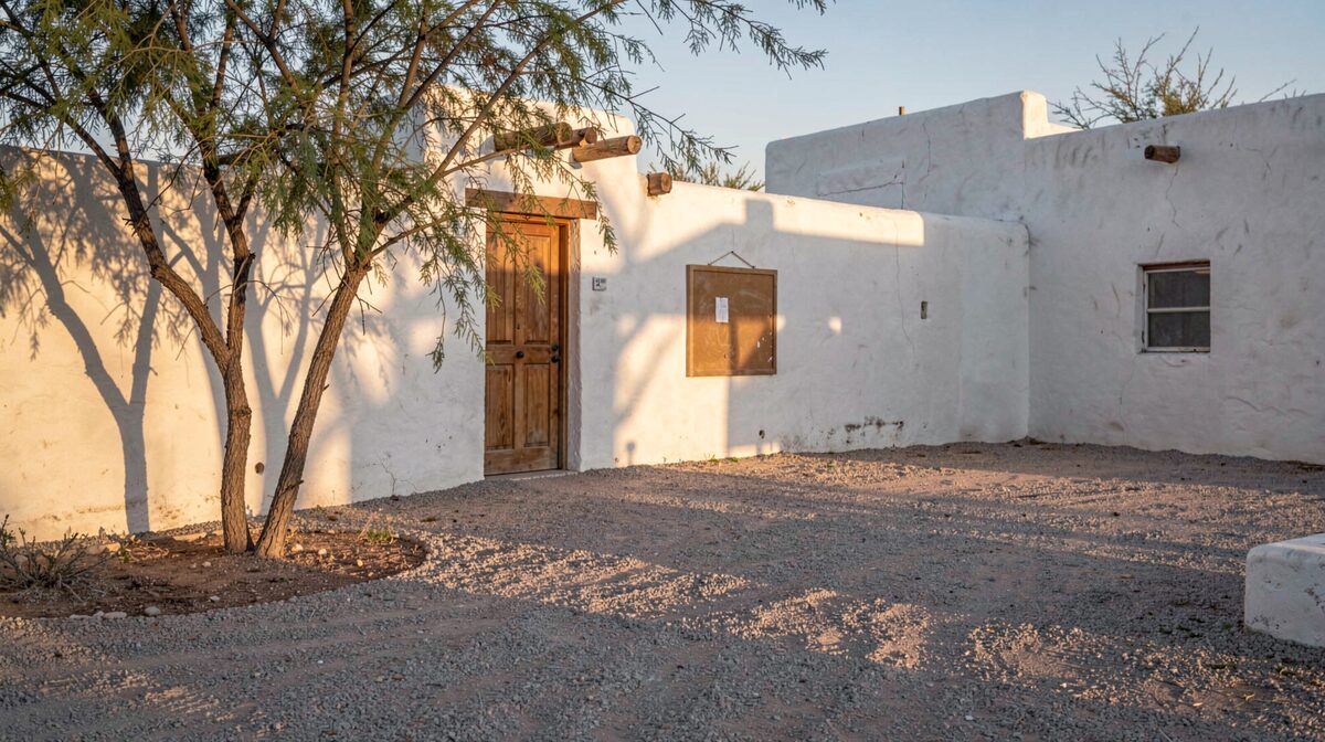 A whitewashed adobe parish wall in low desert light, mesquite branches casting long shadows across a swept gravel path, a plain wooden door and an unlettered notice board, no people and no signage